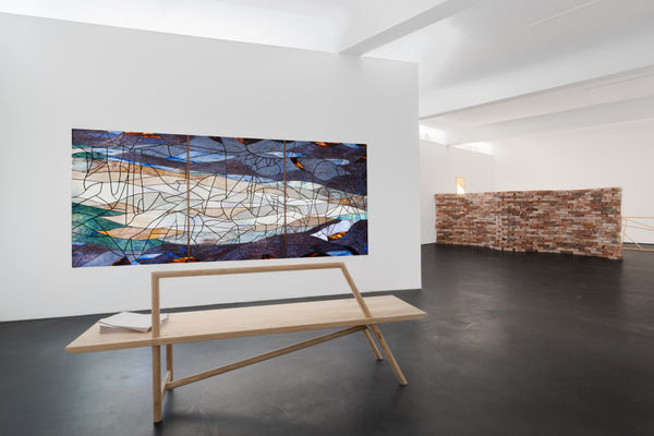 Installation view five | rindon johnson | Photo: Marjorie Brunet Plaza. White room with a dark blue, green and orange stained glass window made up of 3 panels on a plain wall, a red-brown brick wall behind it and a dark floor.