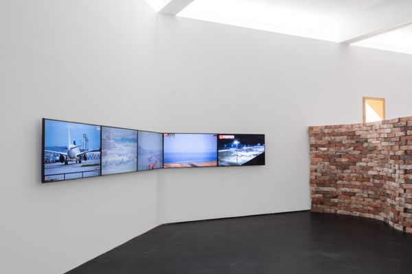 Installation view five | rindon johnson | Photo: Marjorie Brunet Plaza. Wall with five adjacent screens streaming airport scenes, a red-brown brick wall and a window behind it.