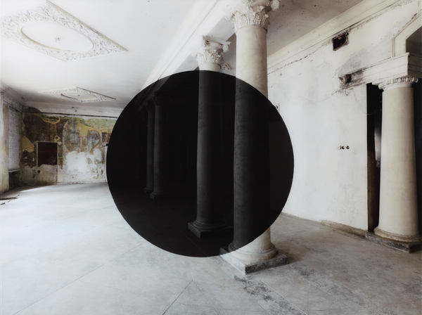 An empty and decaying room with classical columns and a large black circle in the center.