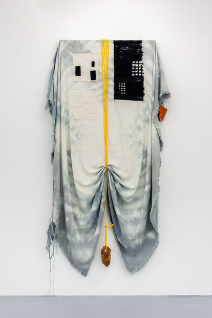 A large sheet of hand died monks cloth hangs from a wooden painting stretcher. The cloth hangs down below the bottom of the stretcher and over both sides. The cloth is sinched at the mid point of the bottom by a nylon rope that wraps around the frame, ove