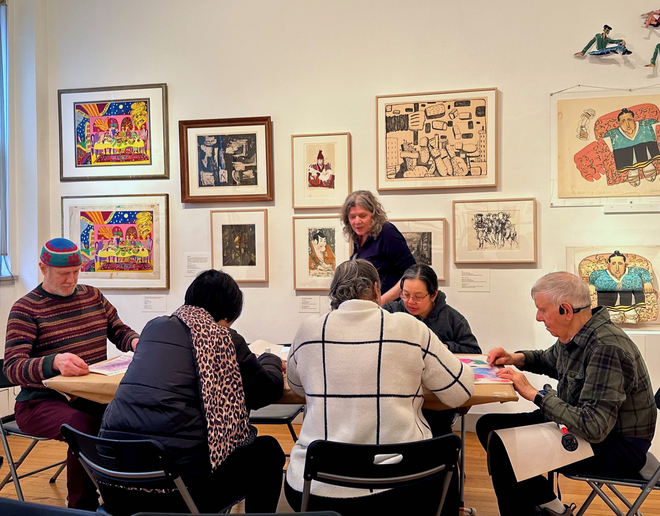 A group of people seated at a table in a gallery with art hanging on the wall behind them. The people seated are looking down at the table while creating art of their own. A woman is standing up, leading the group in the activity.