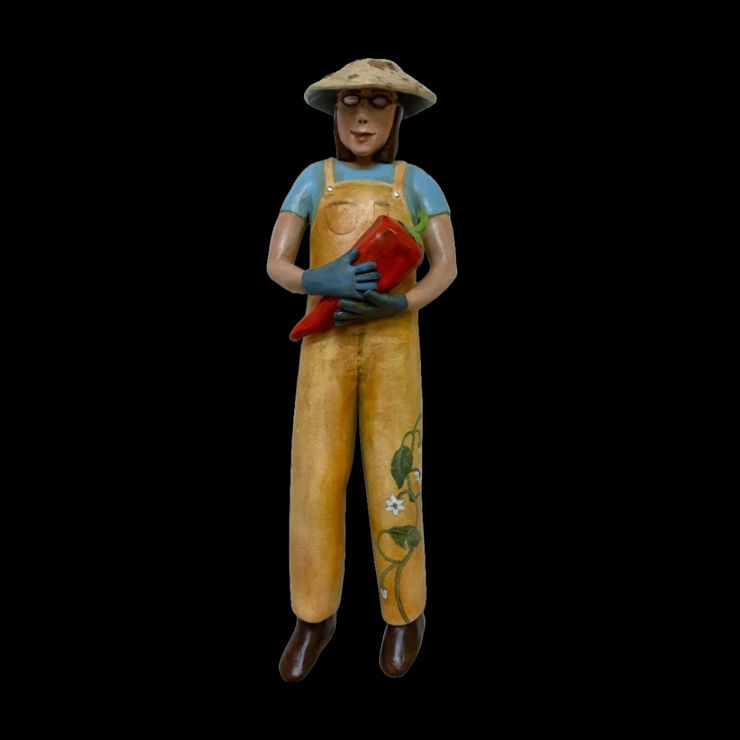 Slate Gray Gallery Artist Julie McNair's clay and oil sculpture of a gardener wearing yellow overalls with a vine on the left leg holding a red pepper titled "pick a pepper"