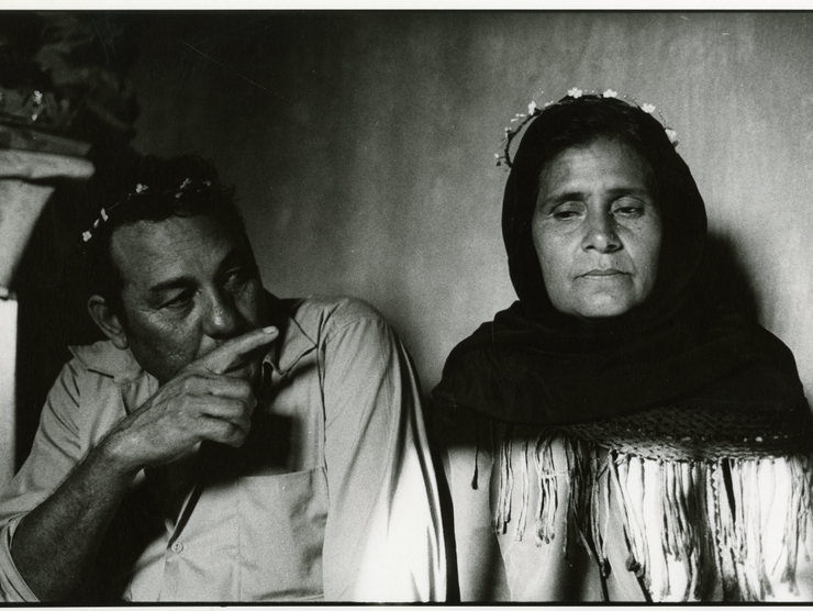A middle-aged man and woman sit next to each other in this black and white photo. She is stoic and he points in her direction.