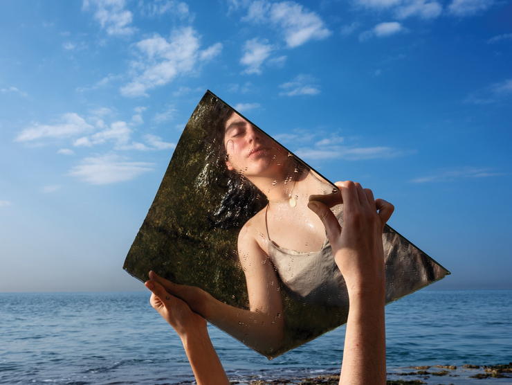 A young woman lays by the ocean and holds up a large piece of mirror showing her reflection.