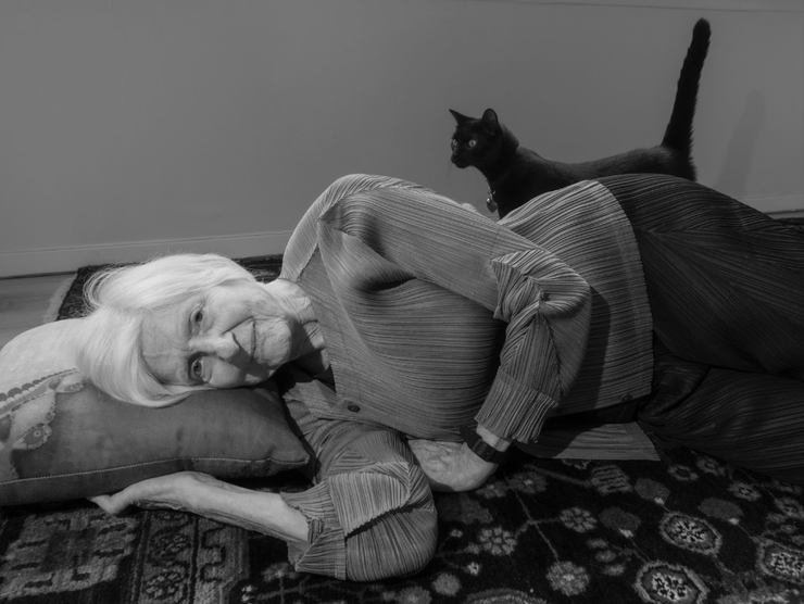 An elderly woman lies down on the floor with a pillow while smiling, with a black cat walking with its tail up behind her.