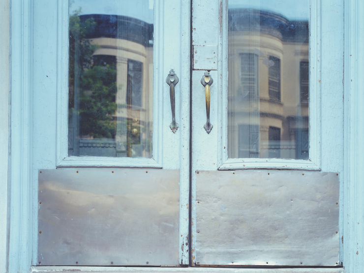 A brownstone is shown reflected in another brownstone's pale blue windowed doorway.