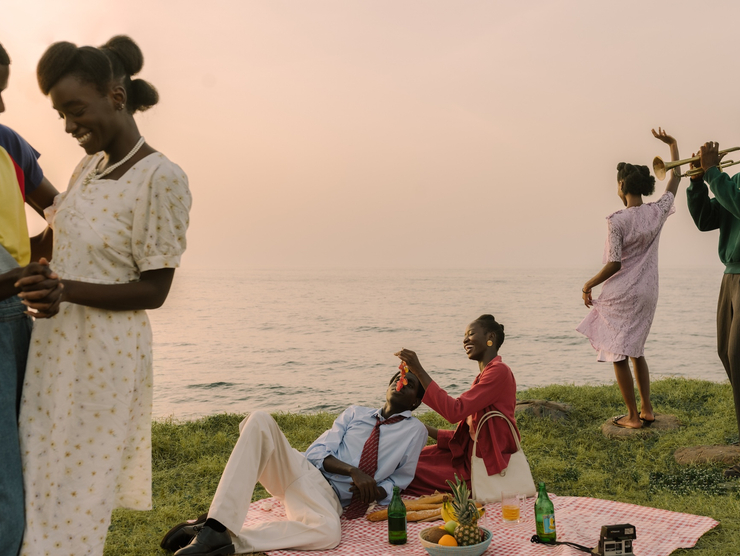 A group of young Black couples have a picnic by a seaside - some dance while smiling, a woman smiles as she feeds grapes to a reclining man, and another man plays the trumpet.