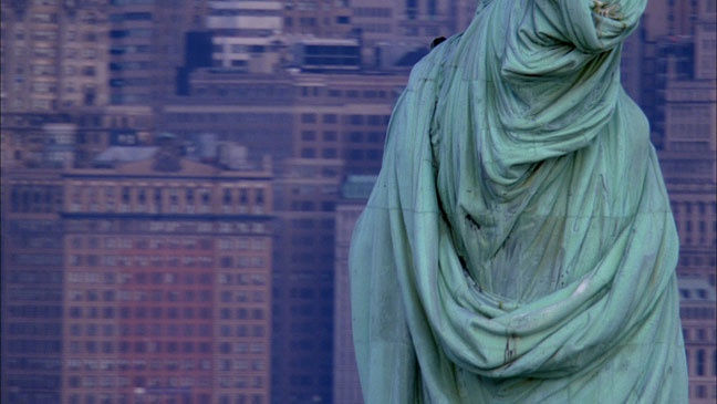 The statue of liberty is cropped at the bust from the posterior view. High-rise buildings fill the background. 