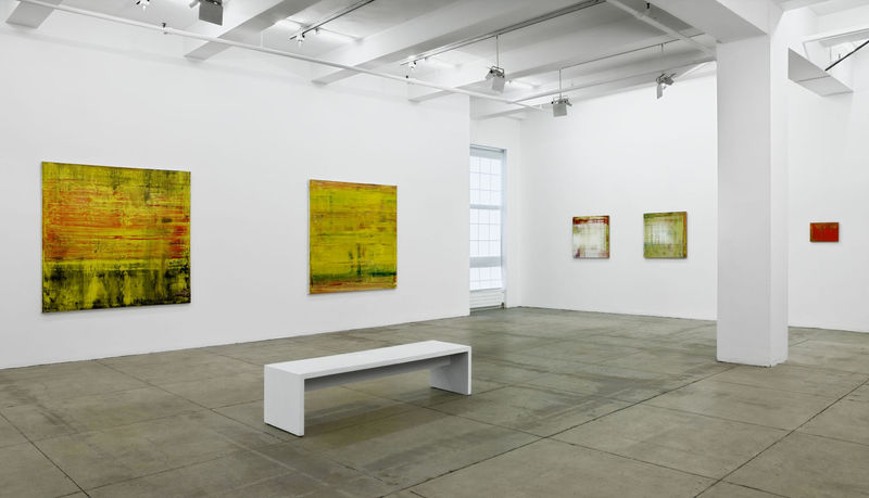 5 abstract paintings of various sizes span two walls with a window in-between. A column and a white bench sit in the center of the room. 