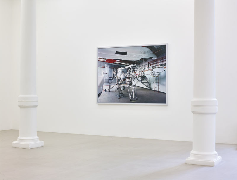 In a white gallery space,  a photograph of an unidentified piece of machinery hangs on a white wall.