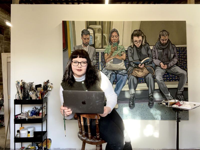 Image shows artist Emily Gillbanks sitting in her studio in front of her painting