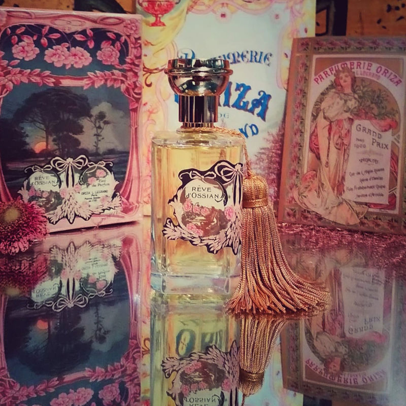 A perfume bottle RÊVE D'OSSIAN by Oriza L. Legrand displayed standing on a mirror with packaging and postal cards in the background