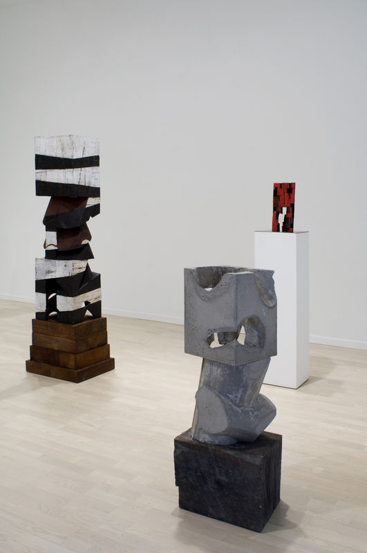 Three Mel Kendrick sculptures on bases and plinths of varied height, the grey on a black squat base, a vertical black and white work, and a smaller red sculpture on the tallest white plinth.