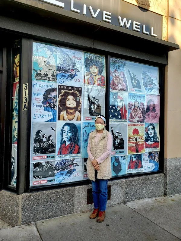 Our founder and owner, Yng-Ru Chen, standing in front of a building with posters plastered over its windows. The building is a GNC store, but the future destination of the gallery. Yng-Ru, the woman standing in front, is wearing a mask.