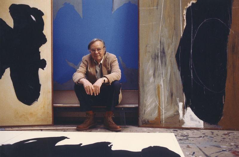 A photo of artist Robert Motherwell sitting in front of one of his paintings in his studio.