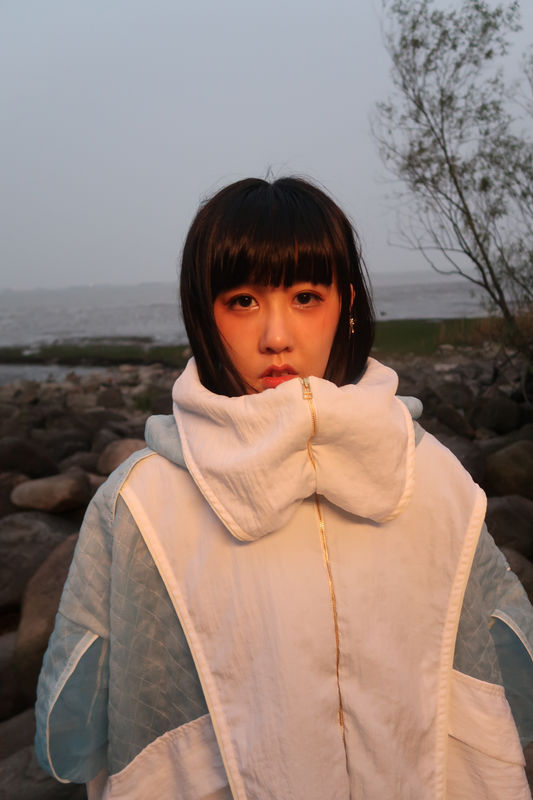 Photograph portrait of artist LuYang outside, wearing a large puffed white and light blue jacket.