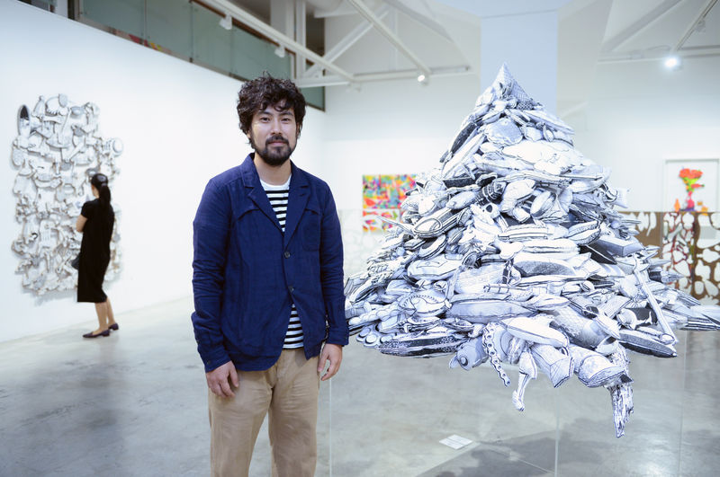 Artist Teppei Kaneuji standing next to one of his soft sculptures inside an exhibition space.