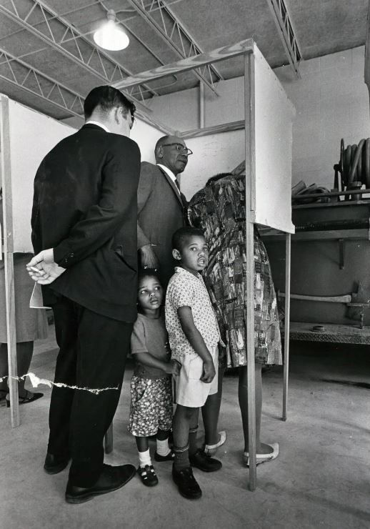 black and white photograph of a woman being assisted in voting by a volunteer worker and her two children in Fayette, Miss., as an election official looks on