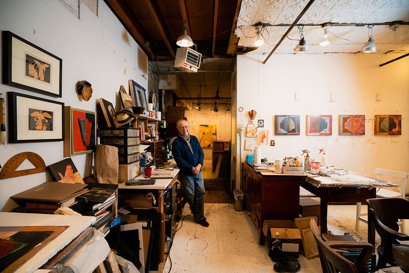 An artist, Joe Haske, leans against a worktable in a loft setting filled with materials and paintings