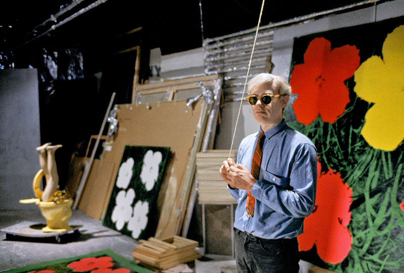 Photograph of Andy Warhol standing in front of his flower paintings, taken by Bob Adelman