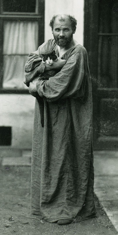 Black and white photograph of the artist Gustav Klimt. He is wearing a flowy robe and holding a cat. He stands looking at the viewer