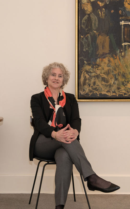 A white woman with curly gray-blond hair wears gray pants, a black blazer and coral scarf as she sits on a chair, hands clasped on her knee, to the left of a gold framed neutral-toned painting on the wall behind her