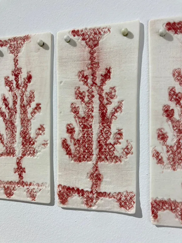 Leena Nammari. Absence Does Not Mean Forgetting. 2026. Seventeen porcelain plaques, emboss printed using a hand-made cross-stitched embroidered tree, glaze-coloured and fired.