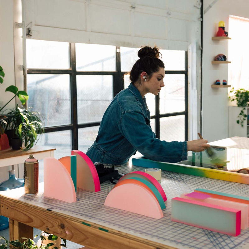 Rachel Strum portrait in studio paintbrush in hand working on painting, neon sculptures in foreground