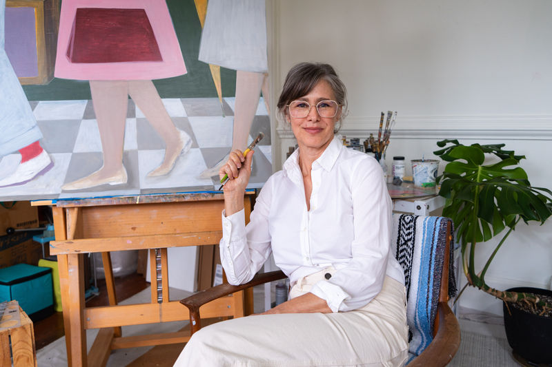 Angela Burson, a middle aged white woman, sits in her light filled studio among her plants and paintings. She is wearing glasses with her hair up and a white shirt