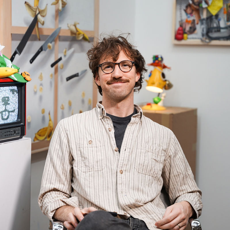 portrait of Stephen Morrison - man with glasses sitting in a chair in front of his artwork (sculpture and paintings)