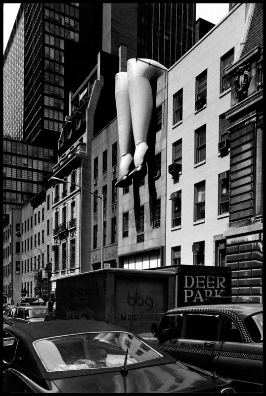 giant blow-up doll's legs in high heels dangling over the side of a building in New York City with cars and trucks on street in foreground by Elliott Erwitt