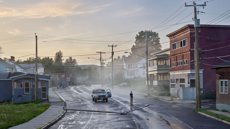 Gregory Crewdson photograph of a misty street with a truck, fallen streetlight, lone figure, and residential buildings at dusk.