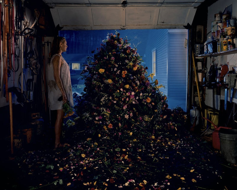 Gregory Crewdson photograph of a woman in a garage at night facing a towering mound of colorful flowers spilling across the floor