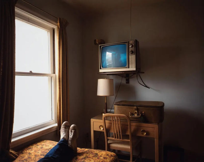 Stephen Shore photograph of a motel room with two beds, patterned bedspreads, a lamp on a nightstand, framed wall art, and daylight through curtained windows.