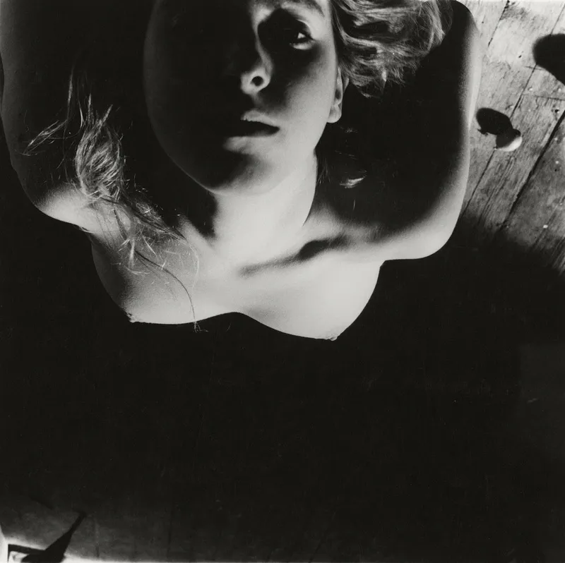Black and white photograph depicting a downward view of an upward-looking nude figure with dramatic lighting.