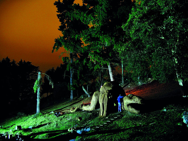 A dinosaur replica laying on a hillside with trees and an orange sky