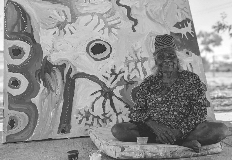 A black and white photo of Aboriginal-Australian artist Tjukupati James sat in-front of one of her paintings.