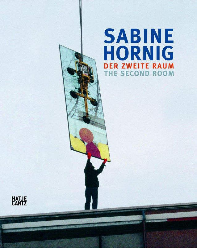 Exhibition catalogue book cover. Photo of a person on a roof holding the bottom of a glass slab sculpture being lowered from a cable above. Text: Sabin Hornig, der Zweite Raum, The Second Room.
