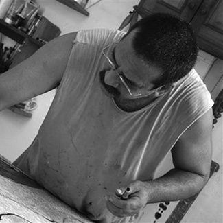 Black and white photograph of a standing over a workbench. He seems to be drawing or painting paper on the bench, just out of view. he wears glasses and has a large, thick, moustache. 