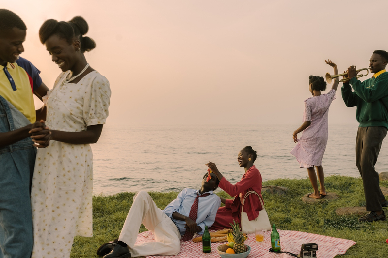 A group of young Black couples have a picnic by a seaside - some dance while smiling, a woman smiles as she feeds grapes to a reclining man, and another man plays the trumpet.