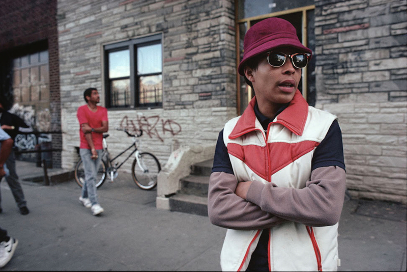 A man on the street in the 1980s wears a maroon hat and stylish jacket with sunglasses and is in the foreground while another man walks by holding his arm. 