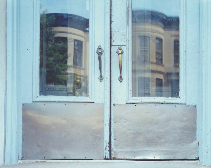 A brownstone is shown reflected in another brownstone's pale blue windowed doorway.