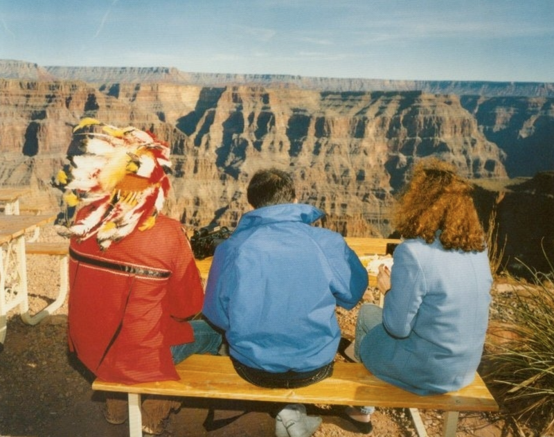 Three people sit on a bench in front of the Grand Canyon, one of them with a Native American feather headress.
