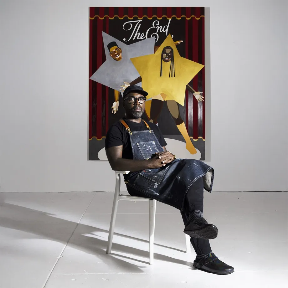 Photograph of Derrick Adams seated in front of a painting that has two faces in the artists painterly style with the text The End above them framed by theatre curtrains