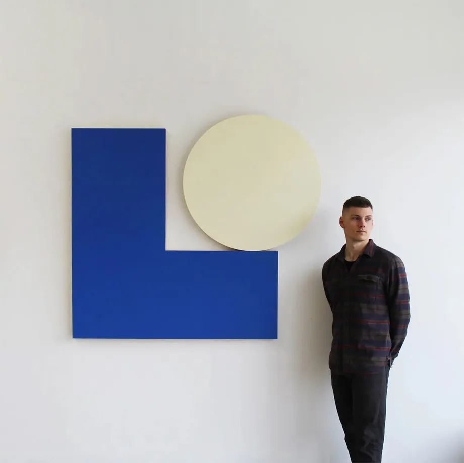 Artist Cameron Ritcher standing next to a bold geometric artwork on the wall comprised of a saturated blue "L" shaped panel and a cream circle.