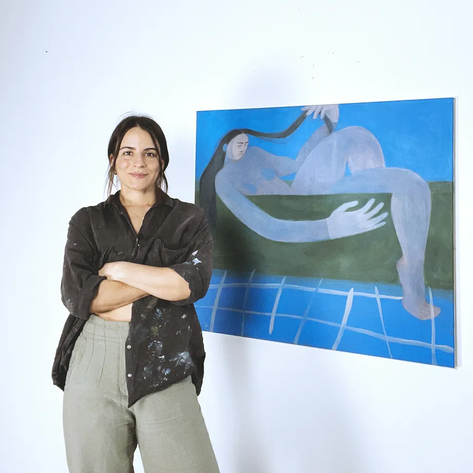 Magdalena Paz, Chilean contemporary painter based in Berlin, standing in her studio beside a large figurative acrylic painting featuring an elongated reclining figure against a blue and green landscape.