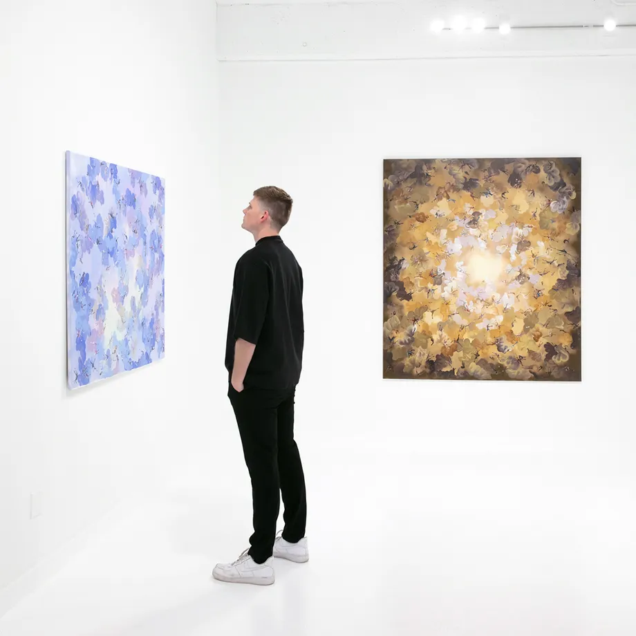 Paul Kuntze Falling Angels exhibition installation view with a person standing in front of two paintings. One with lavender hues featuring abstract human figures amongst aerosol clouds, the other gold and yellow